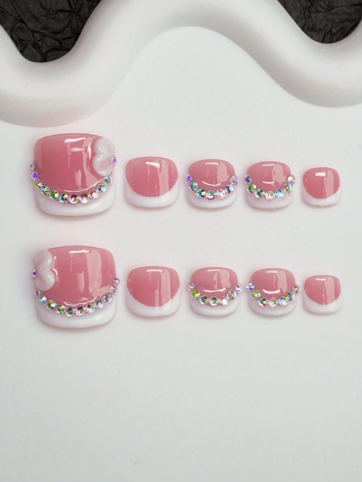 Short square pink French press-on toenails with iridescent crystals and a 3D bow, flat lay